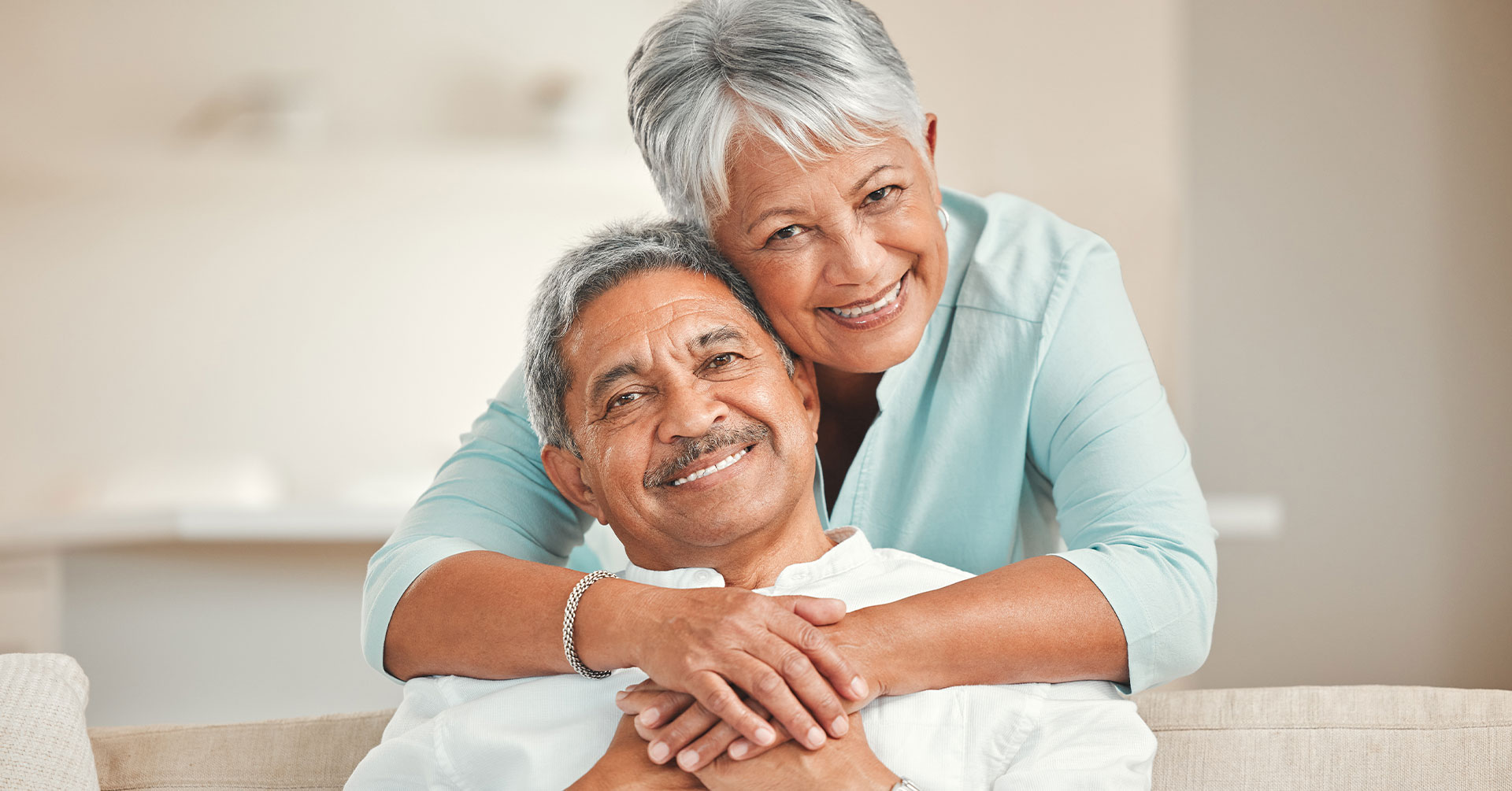 An elderly couple embracing and smiling in a cozy living room setting.
