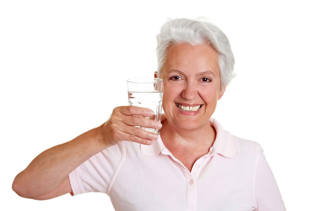 An elderly woman holding a glass of water, smiling and displaying the glass with her right hand.