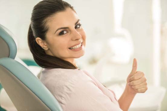 The image shows a woman with dark hair, wearing a blue top and giving a thumbs-up gesture, seated in a dental chair with a smile on her face.