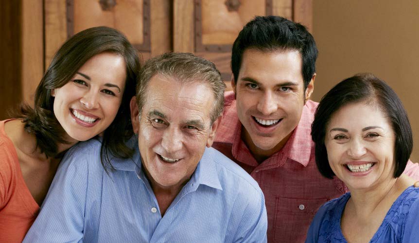 The image is a photograph of four individuals, likely a family, smiling and posing for the camera. They are indoors with warm lighting, and there appears to be a wooden structure or panel behind them.