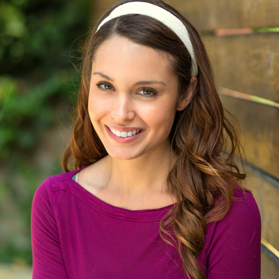 A smiling woman with long brown hair, wearing a purple top and headband, stands in front of a wooden fence.