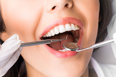 A woman in a white lab coat holding dental instruments near her mouth, with a close-up view of her open mouth.