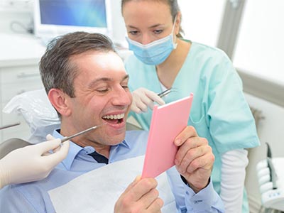 The image shows a man in a dental office, holding up a pink card with a surprised expression, while a female dental professional is smiling and looking at him.