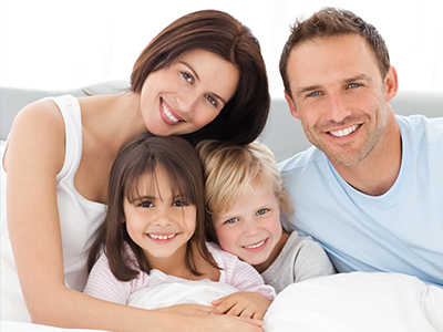 The image shows a family of four - a man, a woman, and two children - smiling and posing together in bed.